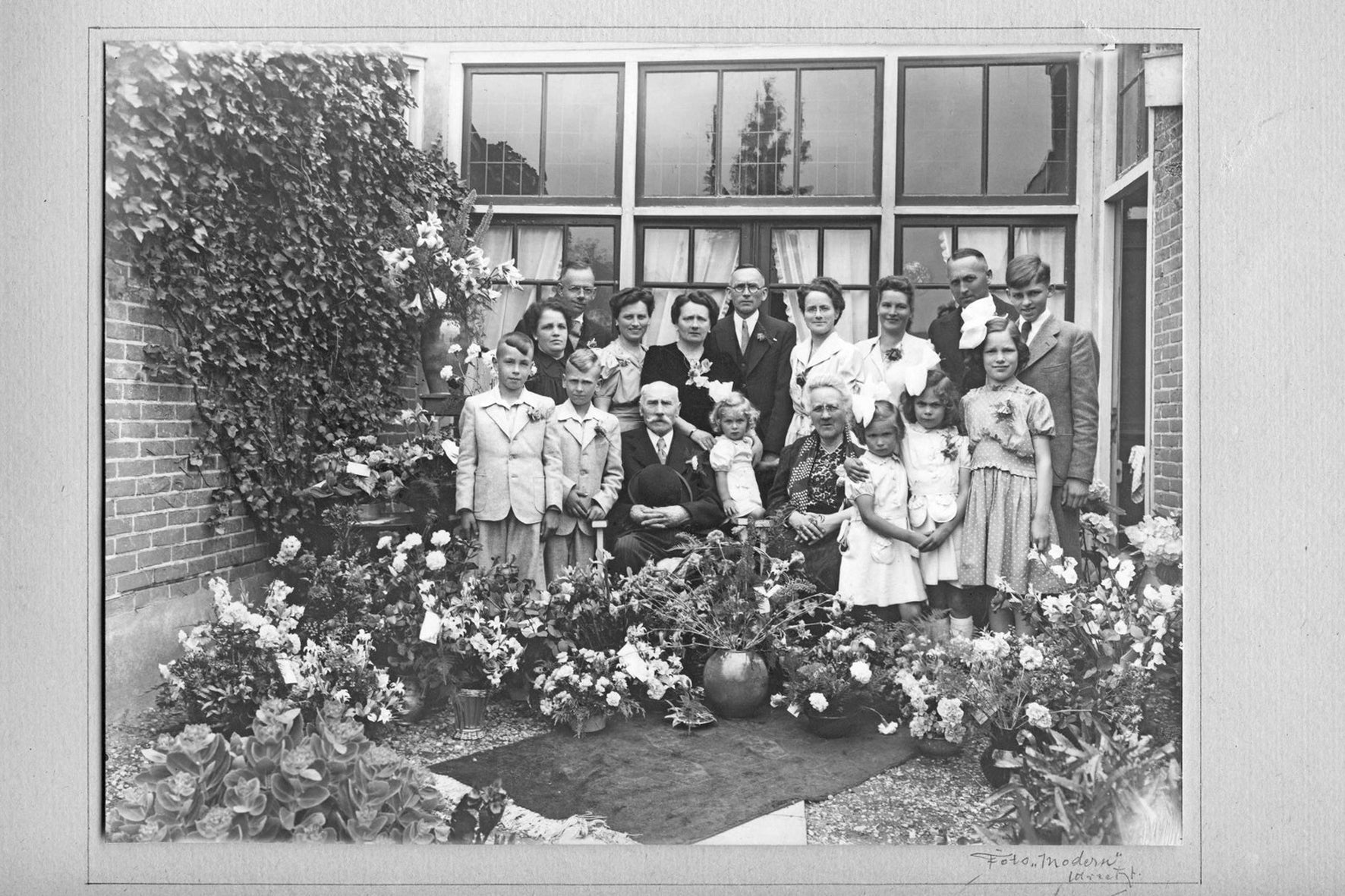A black and white photo of a multi-generational family posing outside in front of a large window. The group includes elderly men and women, middle-aged adults, and children. They are surrounded by numerous potted plants and flowers. Everyone is dressed formally, with the men in suits and women in dresses.