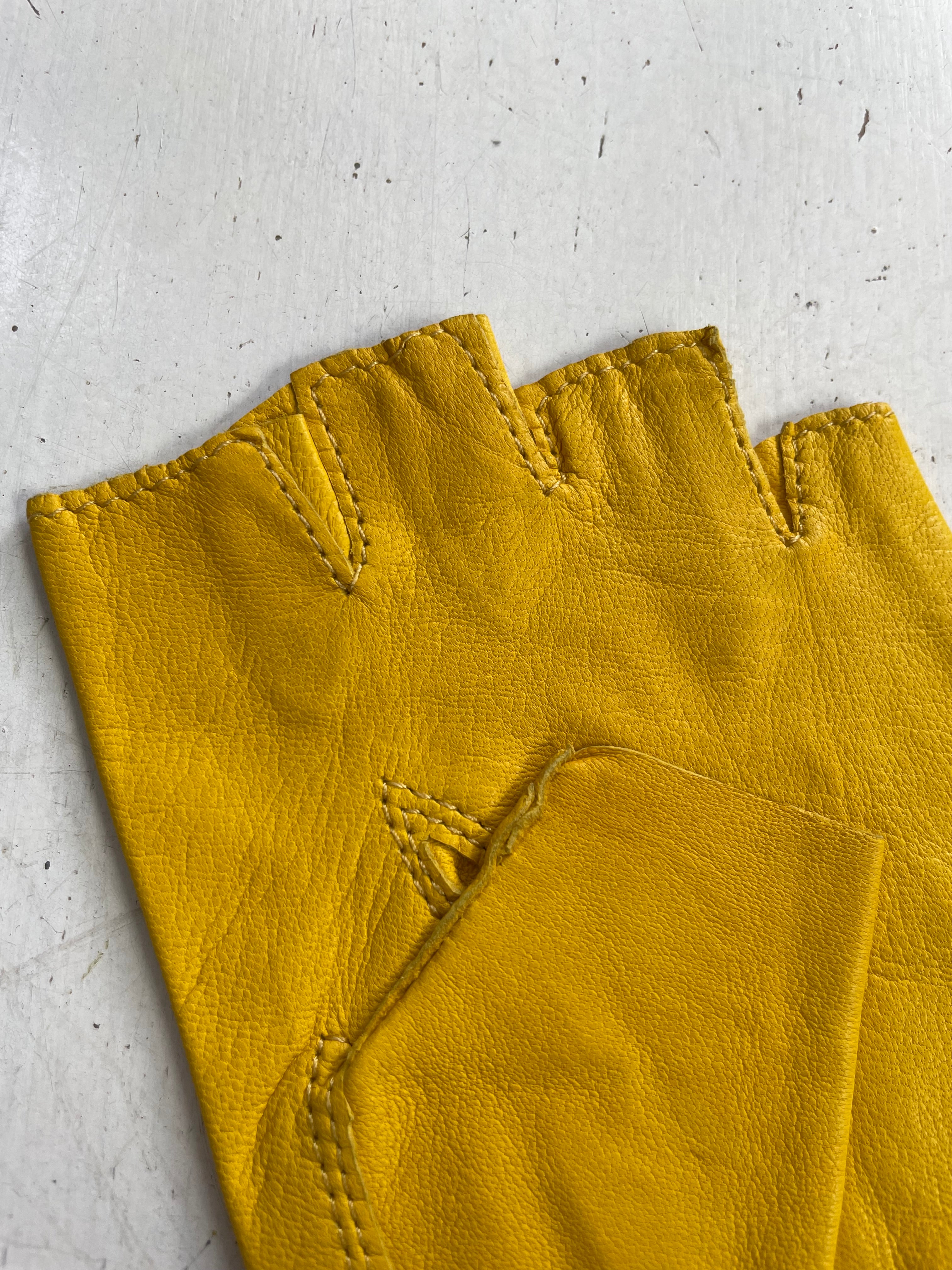 Close-up view of a pair of yellow, deadstock Ines Vintage - Fingerless Fashion Leather Driving Gloves made by Ines Gloves, laying on a white surface. The stitching details are visible, especially around the finger openings and the reinforcement areas on the gloves. The surface underneath is slightly weathered.