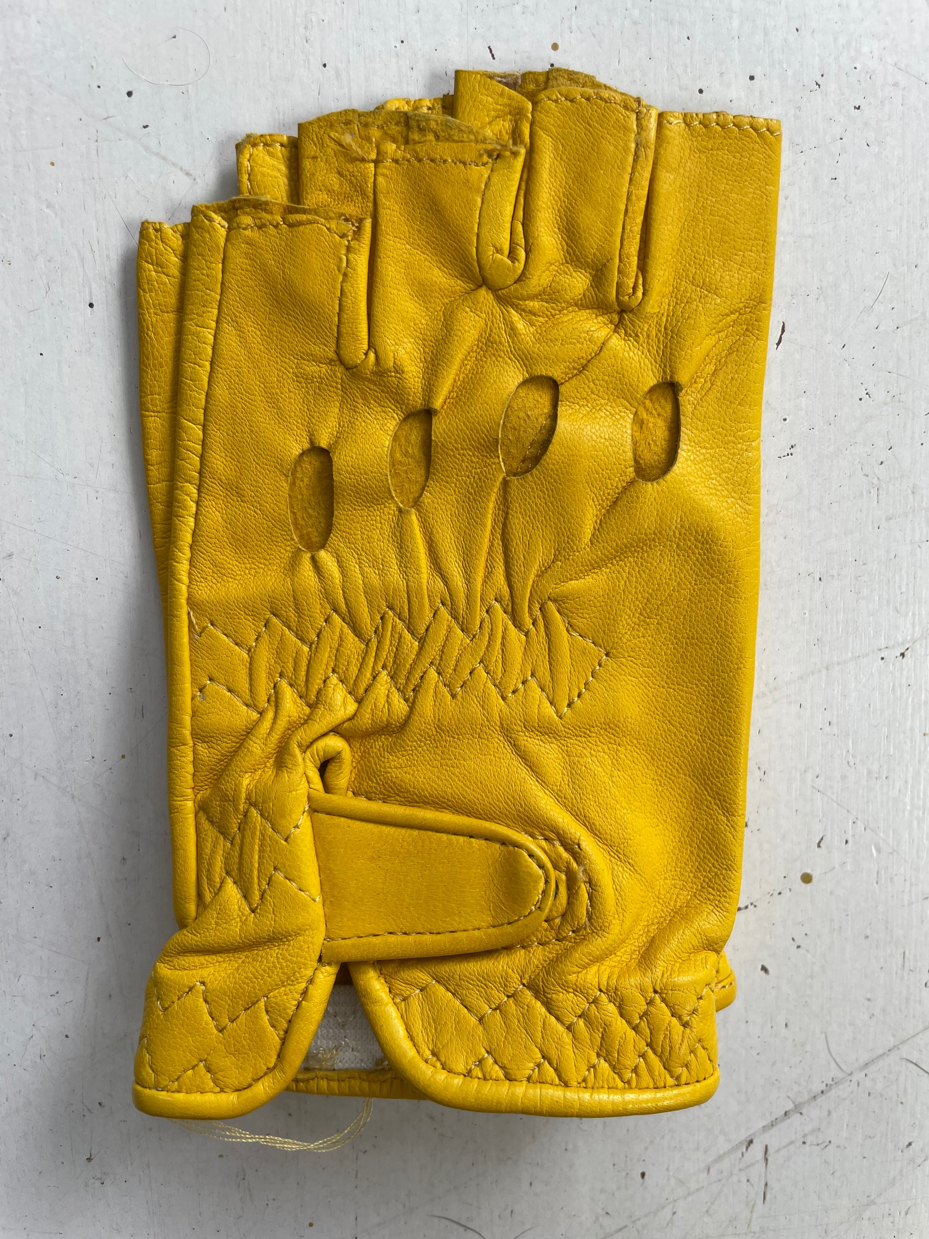 A single yellow leather glove from Ines Gloves' Ines Vintage collection lies on a light-colored surface. This fashionable fingerless driving glove features multiple cut-out sections on the back for ventilation and a strap with Velcro for secure fastening. The textured leather displays visible stitching, particularly around the edges and strap area.