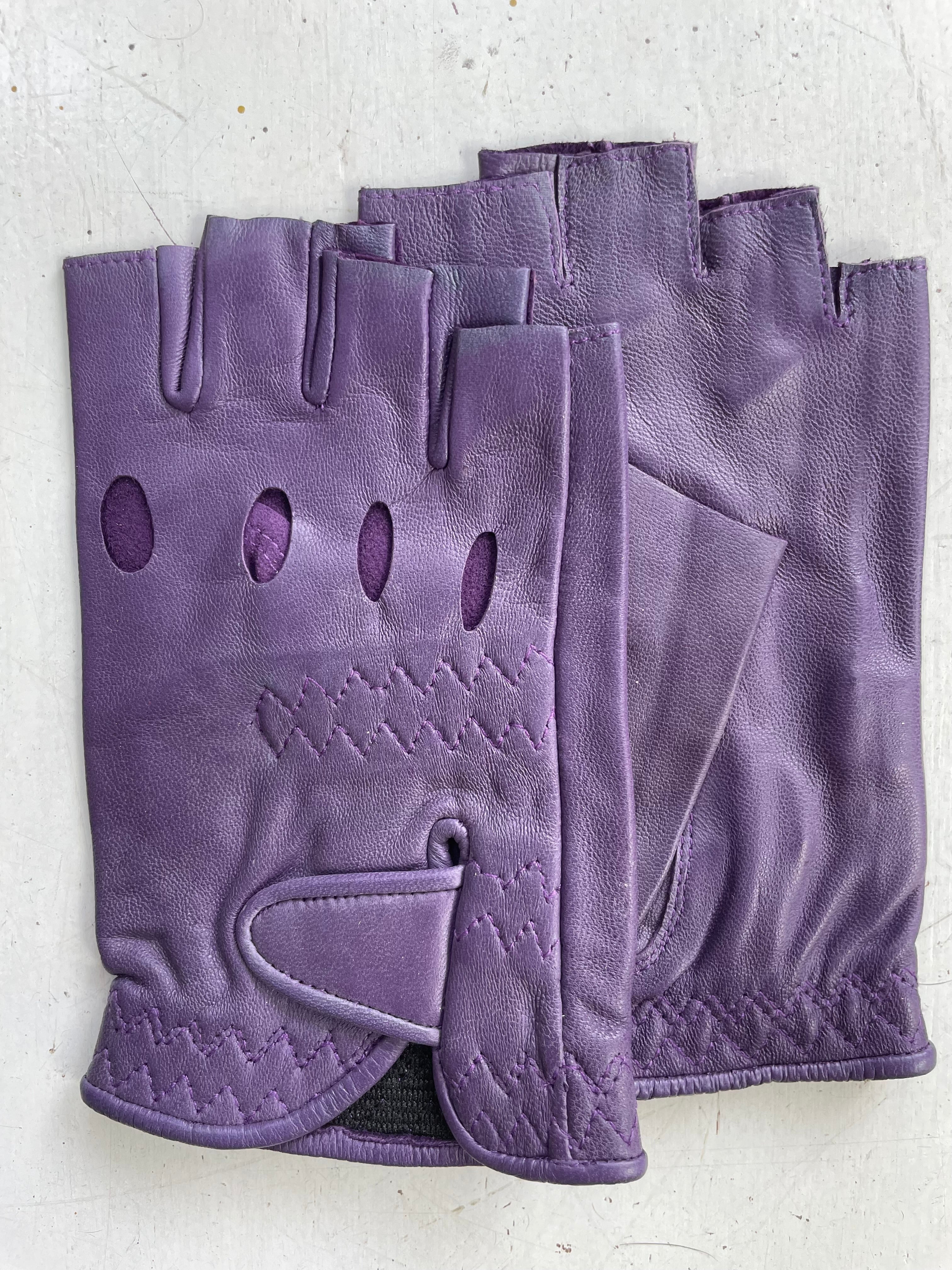 Close-up of a pair of Ines Vintage - Fingerless Fashion Leather Driving Gloves from Ines Gloves, showcased on a white surface. The stylish purple gloves feature decorative stitching and multiple oval perforations along the back for ventilation. An adjustable strap with a Velcro closure is visible on one glove, ensuring a secure fit.