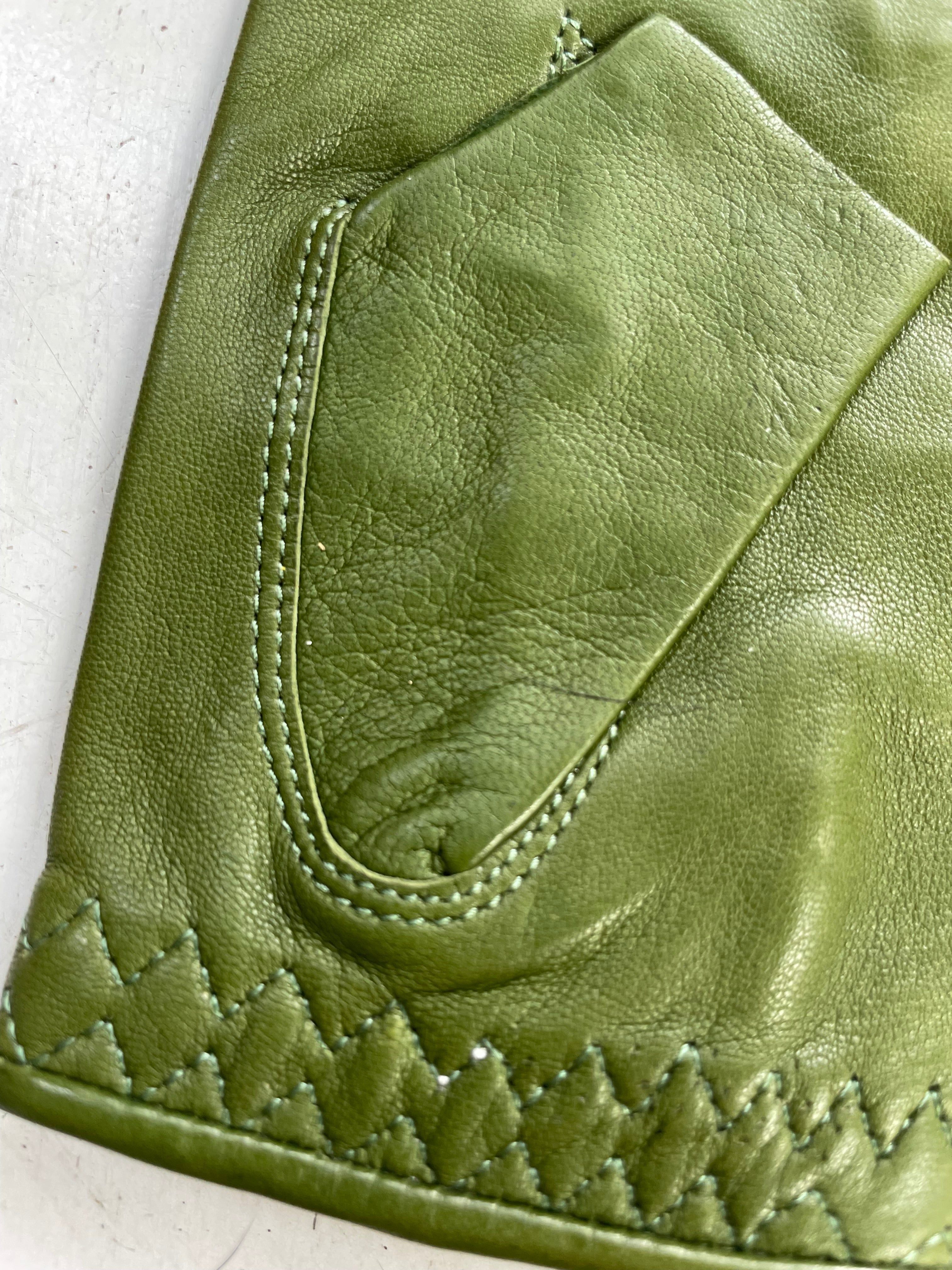 Close-up of the pocket of a green leather garment, showcasing intricate stitching and texture. The pocket features precise, reinforced stitching, and the hemline displays a decorative zigzag pattern. The leather appears soft and supple, hinting at high-quality craftsmanship reminiscent of Ines Gloves Ines Vintage - Fingerless Fashion Leather Driving Gloves.