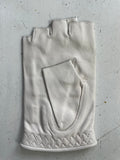A single, white, fingerless leather glove from Ines Gloves is placed on a scratched, light-colored surface. The Ines Vintage - Fingerless Fashion Leather Driving Gloves feature visible stitching and a textured pattern near the wrist area, along with a small pocket on the back of the hand. The surface shows signs of wear with scattered marks and lines.