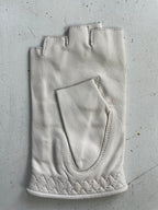A single, white, fingerless leather glove from Ines Gloves is placed on a scratched, light-colored surface. The Ines Vintage - Fingerless Fashion Leather Driving Gloves feature visible stitching and a textured pattern near the wrist area, along with a small pocket on the back of the hand. The surface shows signs of wear with scattered marks and lines.