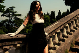 A woman in a black and white dress and long white gloves leans against a stone balustrade with greenery and trees in the background. The scene appears sunlit, highlighting the contrast between her dark dress and the bright backdrop. She gazes off into the distance with a serene expression.