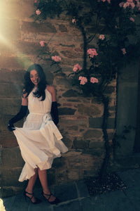 A woman stands against a stone wall adorned with pink flowers and vines. She is wearing a flowing white dress and black gloves, and her black heels complement her outfit. Sunlight filters in from the upper left, casting a warm glow on the scene as she looks down and slightly smiles.