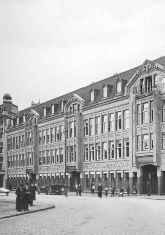 Black-and-white photograph of a street scene featuring a row of ornate, early 20th-century European buildings. The structures have intricate architectural details, large windows, and gabled roofs. Pedestrians and a few cars are visible on the cobblestone street in front of the buildings.