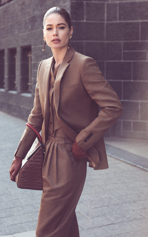 A woman with slicked-back hair walks confidently outdoors, dressed in a stylish brown suit. She accessorizes with elegant earrings and a large textured handbag. Her gloved hands, adorned in classic glacé leather gloves of extra length from Ines Gloves, and composed expression complement the formal outfit. The background features a stone building with evenly spaced windows.