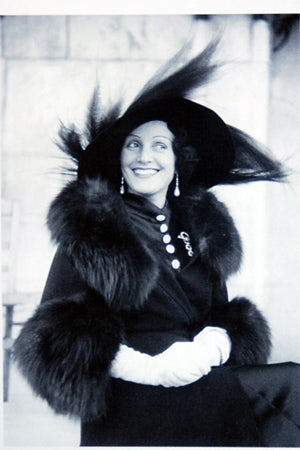 A black-and-white photograph shows a woman dressed in elegant early 20th-century attire. She wears a large hat adorned with feathers, a fur-trimmed cloak, and white gloves. The woman is smiling warmly and looking slightly to her left. The background is blurred with a hint of architectural details.
