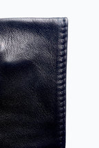 Close-up image of the corner of a pair of black Short fingerless leather fashion gloves by Ines Gloves against a white background. The leather is polished and smooth, with visible texture and stitching along the edge, reminiscent of handcrafted gloves. The stitching is precisely done, indicating quality craftsmanship. The gloves are folded, showing their thickness.