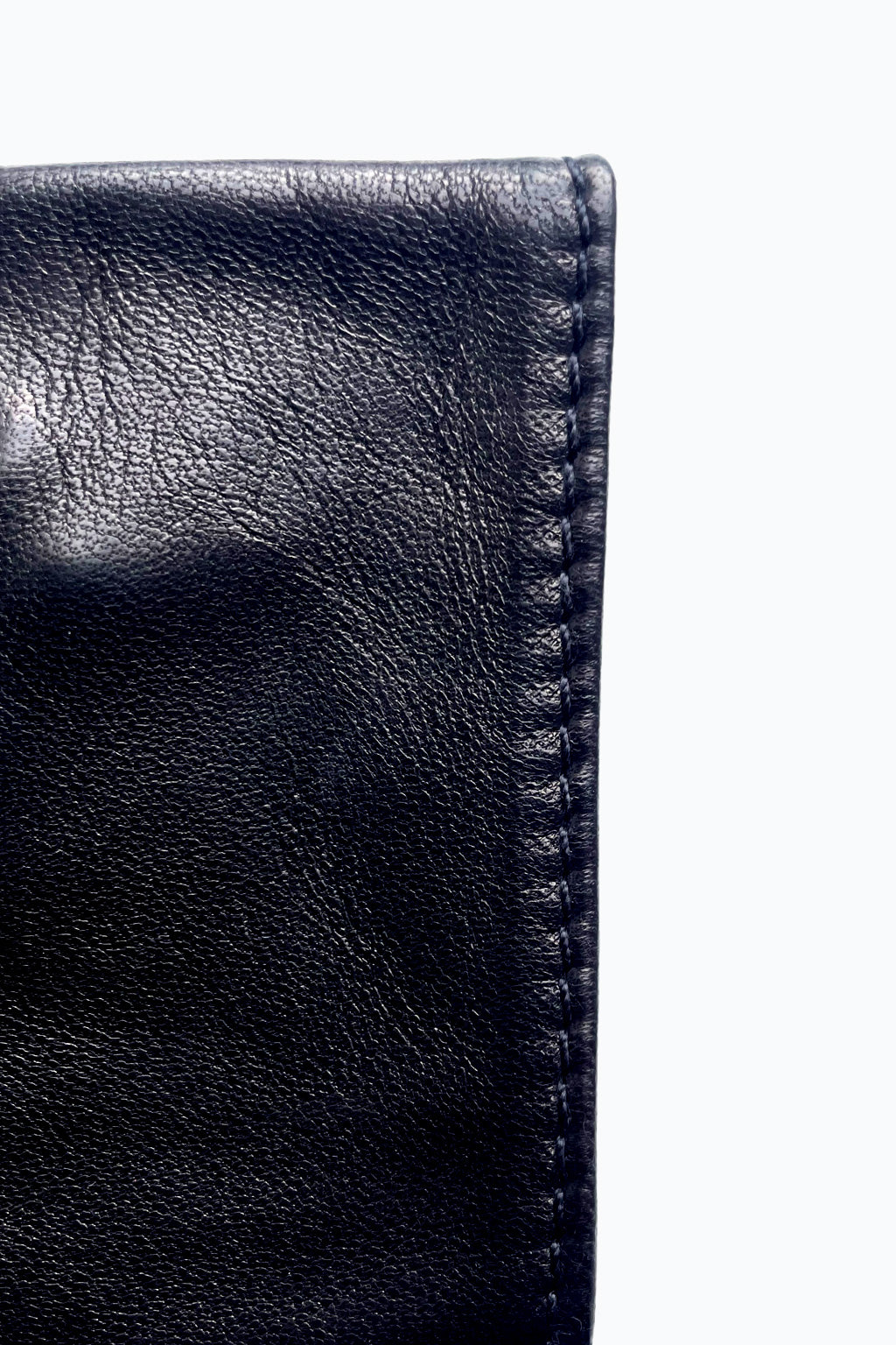 Close-up image of the corner of a pair of black Short fingerless leather fashion gloves by Ines Gloves against a white background. The leather is polished and smooth, with visible texture and stitching along the edge, reminiscent of handcrafted gloves. The stitching is precisely done, indicating quality craftsmanship. The gloves are folded, showing their thickness.