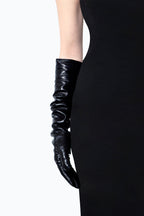 A person wearing a form-fitting, sleeveless black dress is shown from the shoulders to the hips, with their right arm extended downward. The person is also wearing long cashmere lined gloves by Ines Gloves that extend past the elbow, adding a sophisticated touch to the outfit.