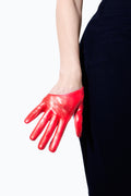 A person wearing an Ines Gloves Half Scoop Leather Glove on their left hand rests it against their hip. The hand is relaxed and the glove contrasts sharply with the dark blue clothing they are wearing. This striking image recalls an iconic scene from Sex and the City, with the red glove standing out prominently against the white background.