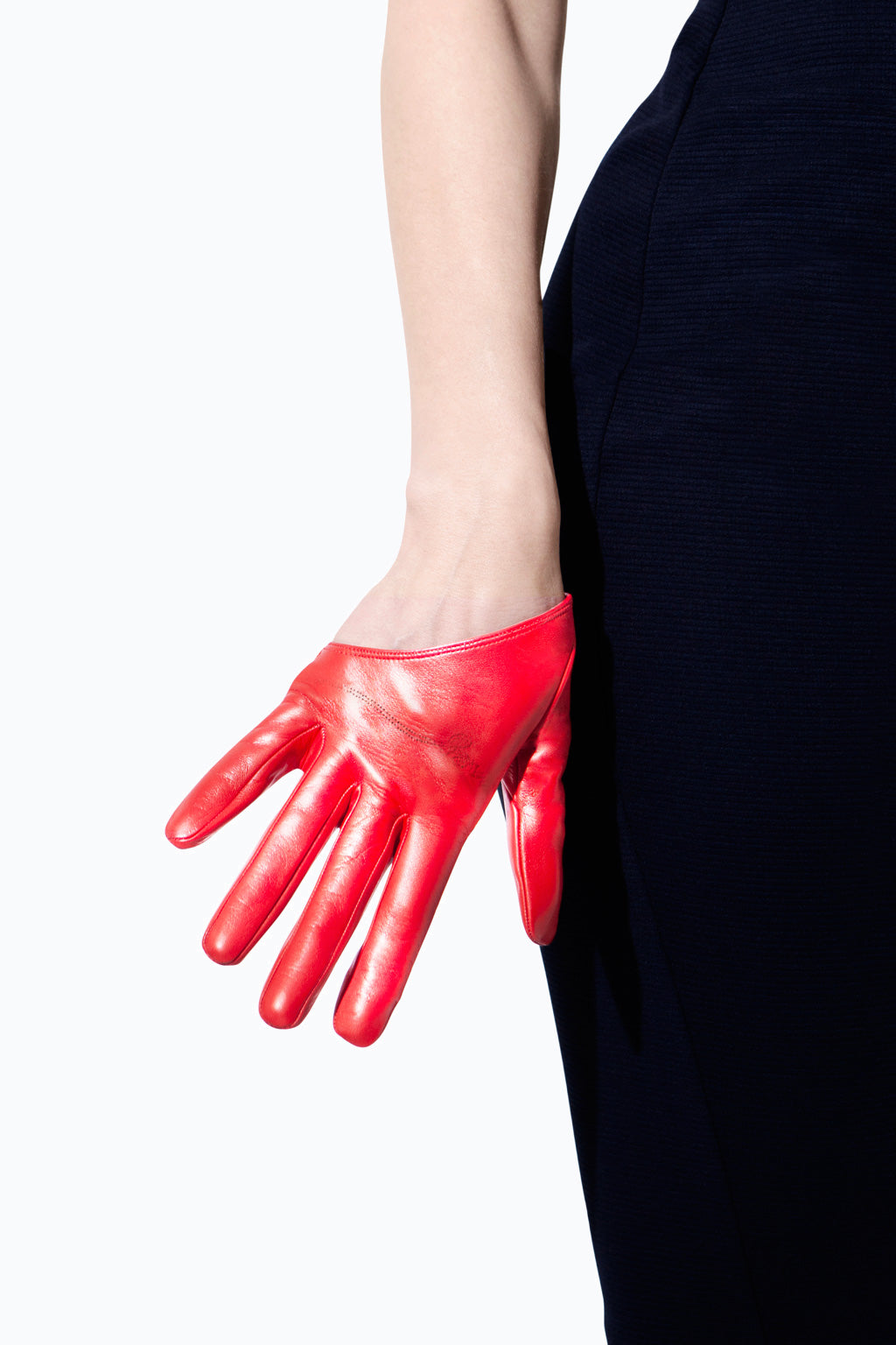 A person wearing an Ines Gloves Half Scoop Leather Glove on their left hand rests it against their hip. The hand is relaxed and the glove contrasts sharply with the dark blue clothing they are wearing. This striking image recalls an iconic scene from Sex and the City, with the red glove standing out prominently against the white background.