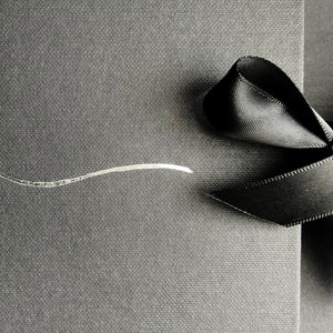 A close-up image of black textured paper, partially covered by a smooth, glossy black ribbon tied into a simple loop. A thin, slightly curved, silver metallic line decorates the left side of the paper, adding a subtle contrast to the dark, elegant background—a perfect setting for an Ines Gloves Gift Card from Ines Gloves.