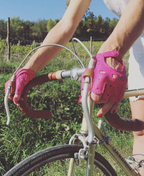A person wearing pink Fingerless Peccary Driving Gloves by Ines Gloves grips the handlebars of a road bicycle. The bike features tan Peccary leather handles and brake cables. The cyclist is dressed in a white sleeveless top and white bottoms. A scenic background of green foliage, trees, and a sunny sky is visible.