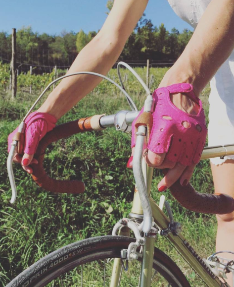 A person wearing pink Fingerless Peccary Driving Gloves by Ines Gloves grips the handlebars of a road bicycle. The bike features tan Peccary leather handles and brake cables. The cyclist is dressed in a white sleeveless top and white bottoms. A scenic background of green foliage, trees, and a sunny sky is visible.