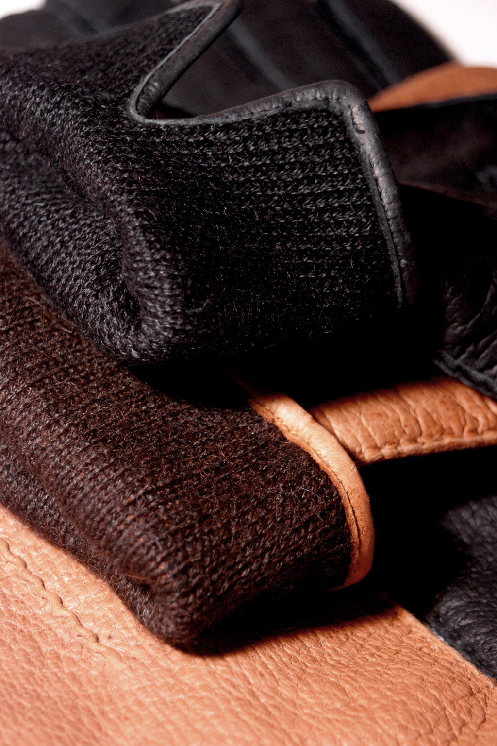 Close-up image of a stack of gloves. The top glove is black with a fabric texture, the middle glove is genuine peccary leather, and the bottom glove is brown peccary leather. The image focuses on the different materials and textures of the Alpaca lined peccary leather gloves by Ines Gloves, highlighting the contrast between fabric and leather.