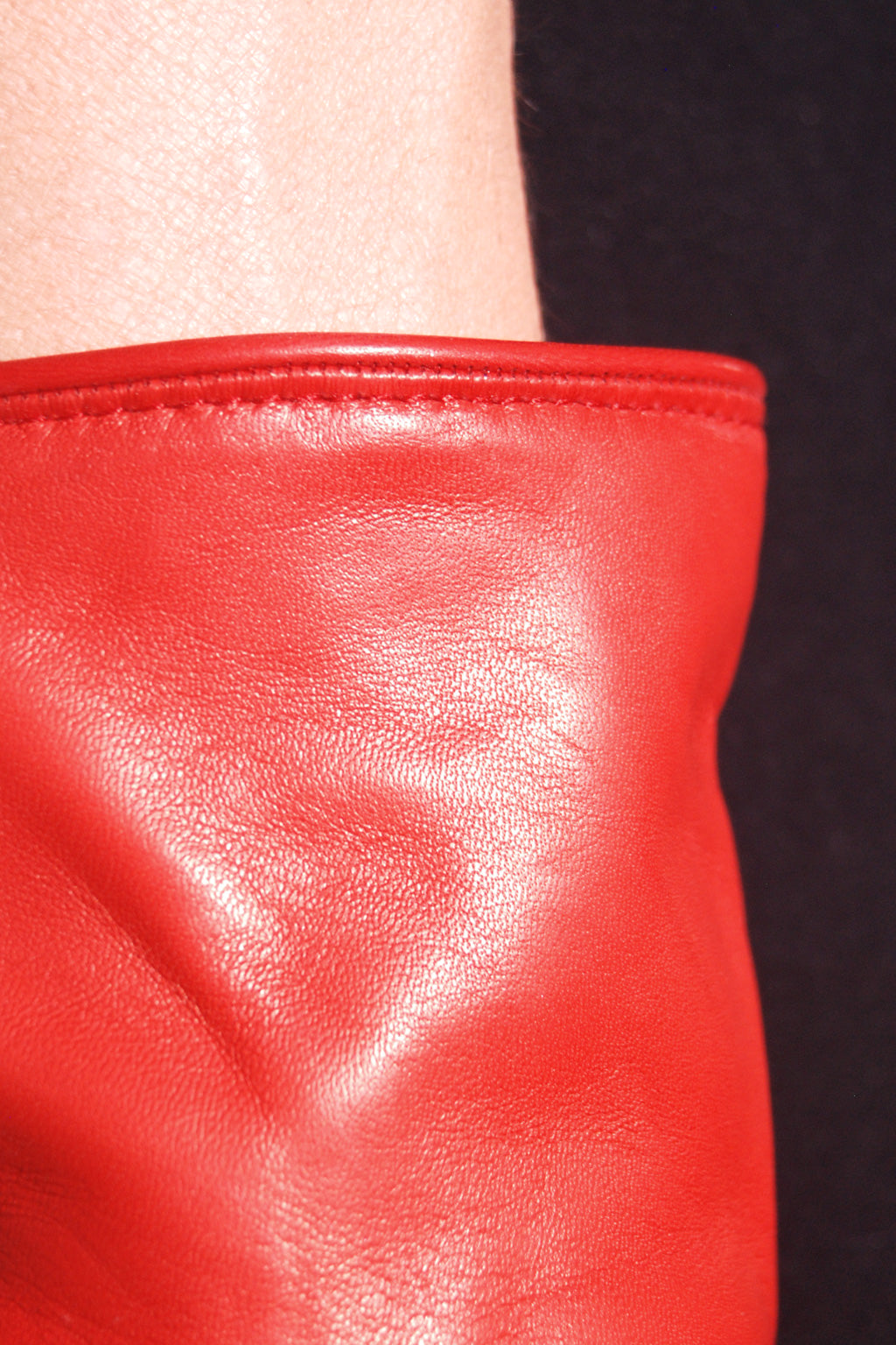 Close-up of a pair of Ines Gloves long cashmere lined gloves, likely a sleeve or long glove, against a dark background. The texture of the leather is smooth with visible creases and a shiny finish. The top edge of the garment has a neatly stitched seam, and a beige area, possibly a person's arm, is visible at the top.