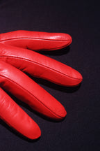A close-up image of **Ines Gloves Long cashmere lined gloves** placed on a dark, possibly black surface. The lighting accentuates the texture and seam details of the gloves, revealing their luxurious cashmere lining. Only the fingers of the glove are visible, fanned out slightly. The contrast between the vibrant red and the dark background is striking.
