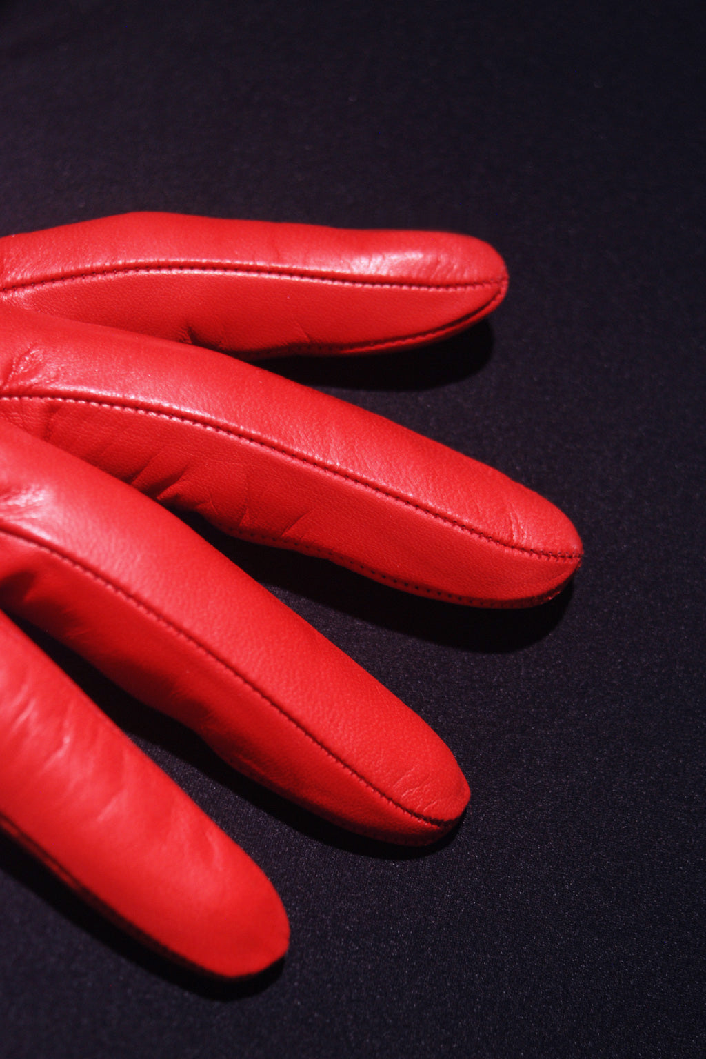 A close-up image of four fingers of a bright red handcrafted Classic cashmere lined leather gloves by Ines Gloves against a dark background. The gloves have visible stitching along the edges, and the texture of the leather is smooth with slight wrinkles. The lighting highlights the rich color and detail of the glove's material.