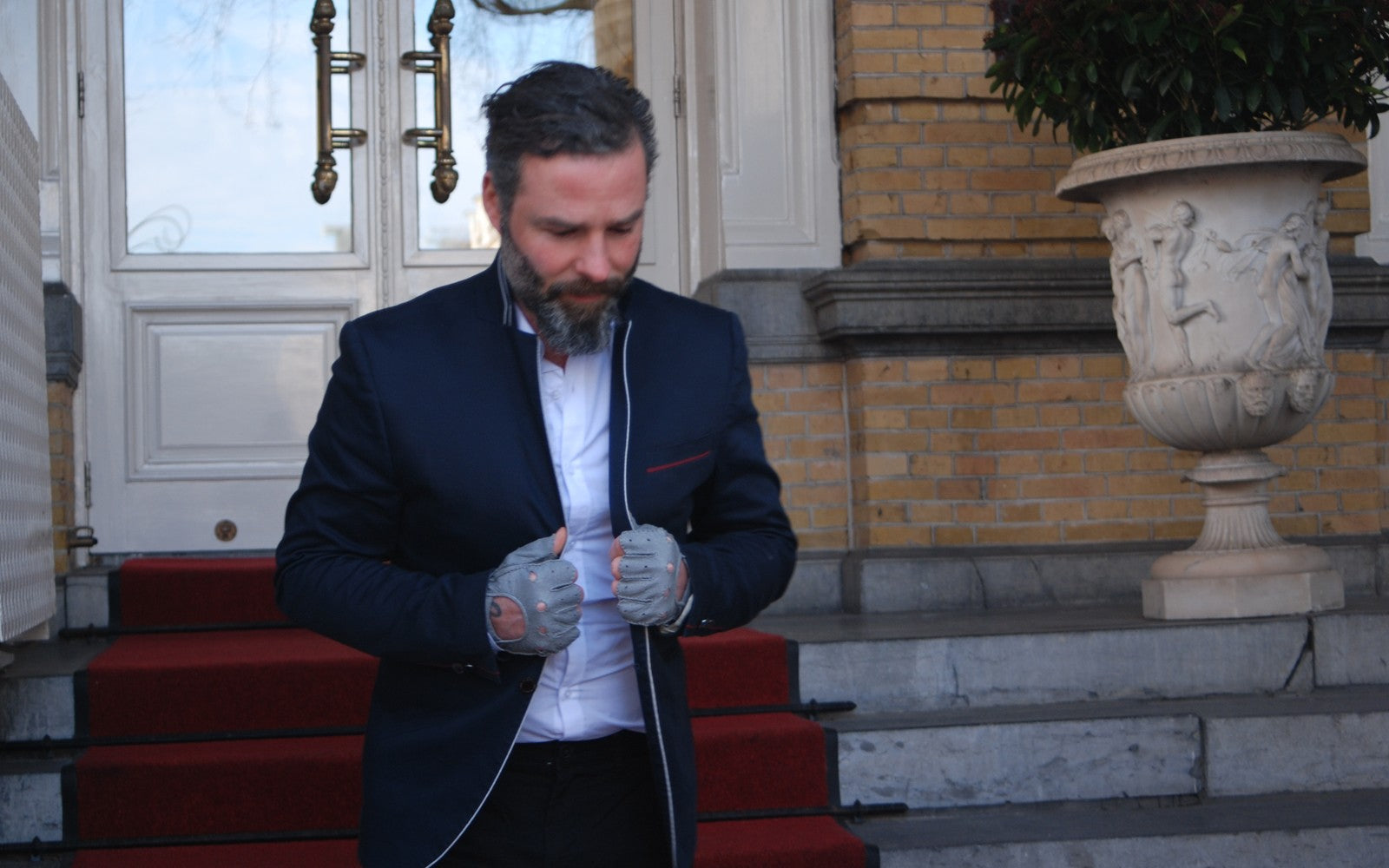A man with a beard, wearing a navy blazer, zips up his jacket while standing on stairs with red carpeting. He is in front of a building with yellow brick walls, ornate white doors, and a large decorative planter. He is also wearing Ines Gloves fingerless driving peccary gloves.